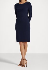 Marinblå åtsittande klänning med långa ärmar, rund halsringning, guldknappaccent på axlarna, slät textur, knäkort. Bruna remsandaler med hög klack.