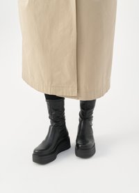 Botas de cuero negras hasta la rodilla con una suela de plataforma gruesa; textura suave con ligero brillo; combinadas con un abrigo beige.