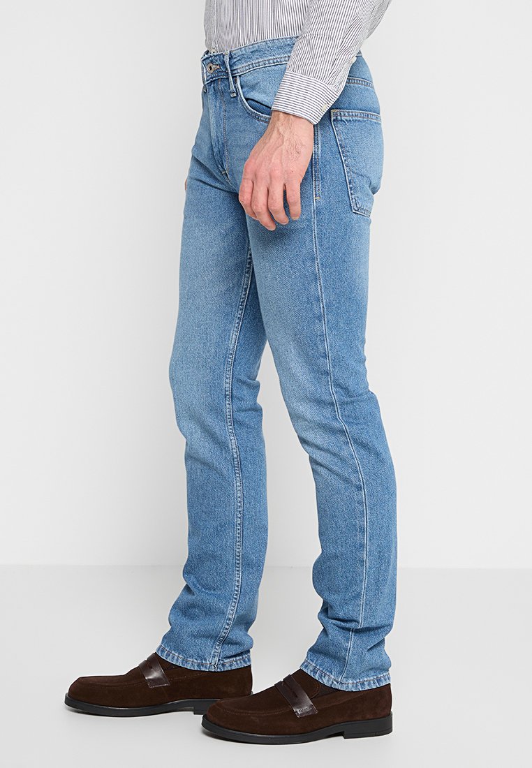 Hombre vestido con jeans de corte slim de color azul claro, mocasines de ante marrones y una camisa de rayas metida por dentro, de pie frente a un fondo blanco liso.
