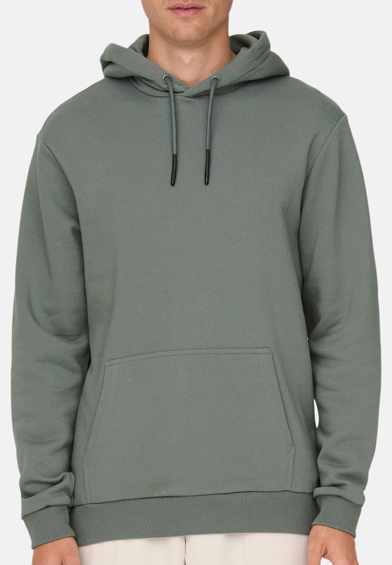 Uomo che indossa una felpa con cappuccio verde tenue, senza disegni, con tasca frontale a sacco e cappuccio con coulisse, mostrato dalle spalle fino ai fianchi.