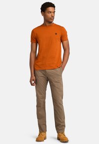 Orange bomulds T-shirt med rund halsudskæring og lille logo på brystet, parret med beige bukser og brune arbejdssko.