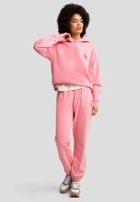 Haut à capuche rose clair avec un design court, des poignets côtelés et un petit logo rouge ; pantalon de survêtement rose assorti avec des poignets élastiques aux chevilles.
