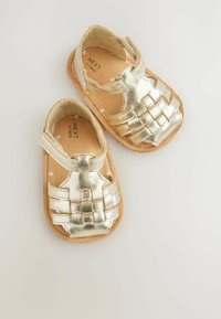 Gold-colored infant sandals with a woven design, featuring a Velcro strap, fabric lining, and a cushioned cork footbed.