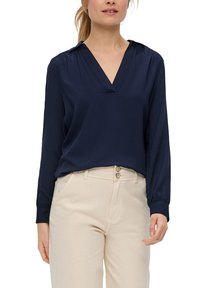 Marineblaue Bluse mit langen Ärmeln und V-Ausschnitt, mit einer glatten Textur und einem dezenten Kragen. Kombiniert mit hellbeigen Hosen mit doppelten Knöpfen.