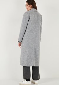 Grijze wollen jas met een lange silhouette, notch-kraag en zijsplitten. Gecombineerd met donkere jeans en witte sneakers, wat een casual uitstraling geeft.