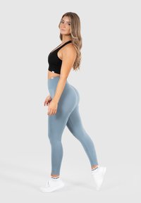 Zwarte sportbeha met een open rugontwerp, gecombineerd met een aansluitende lichtblauwe legging, die een gladde textuur en hoge tailleband toont. Witte sneakers.