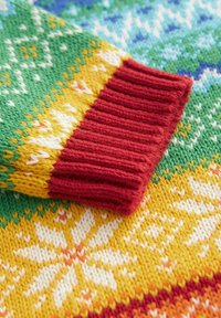 Detalle de un suéter de punto colorido que presenta un puño acanalado en rojo brillante, intrincados patrones florales en blanco y naranja, sobre fondos verdes y amarillos.