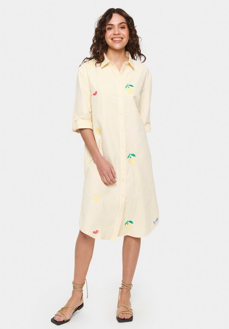 Vestito camicia giallo chiaro con righe verticali. Presenta disegni di frutta ricamati in rosa, verde e blu. Maniche lunghe arrotolate.