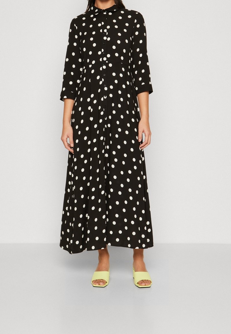 Femme portant une robe noire à manches longues boutonnée avec des pois blancs et des sandales à talons ouvertes jaunes.