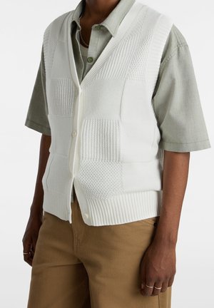 Personne portant un pantalon beige, une chemise verte à manches courtes boutonnée, et un gilet tricoté blanc texturé avec boutons et ourlet côtelé.