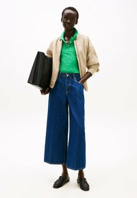 Giacca bomber beige, polo verde, jeans blu a gamba larga e mocassini neri. Modello che tiene una borsa a tracolla nera texturizzata.