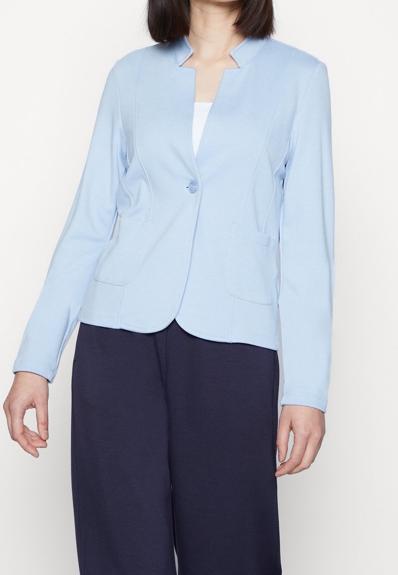 Femme portant un blazer bleu clair à un bouton avec poches plaquées sur un haut blanc et un pantalon foncé à jambes larges, debout devant un fond uni.