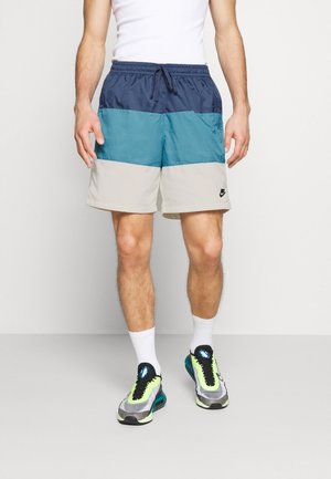 Pantaloncini a blocchi di colore in tessuto blu navy, verde petrolio e beige, con vita elasticizzata e coulisse. Presentano un logo nero sulla parte bassa della gamba.