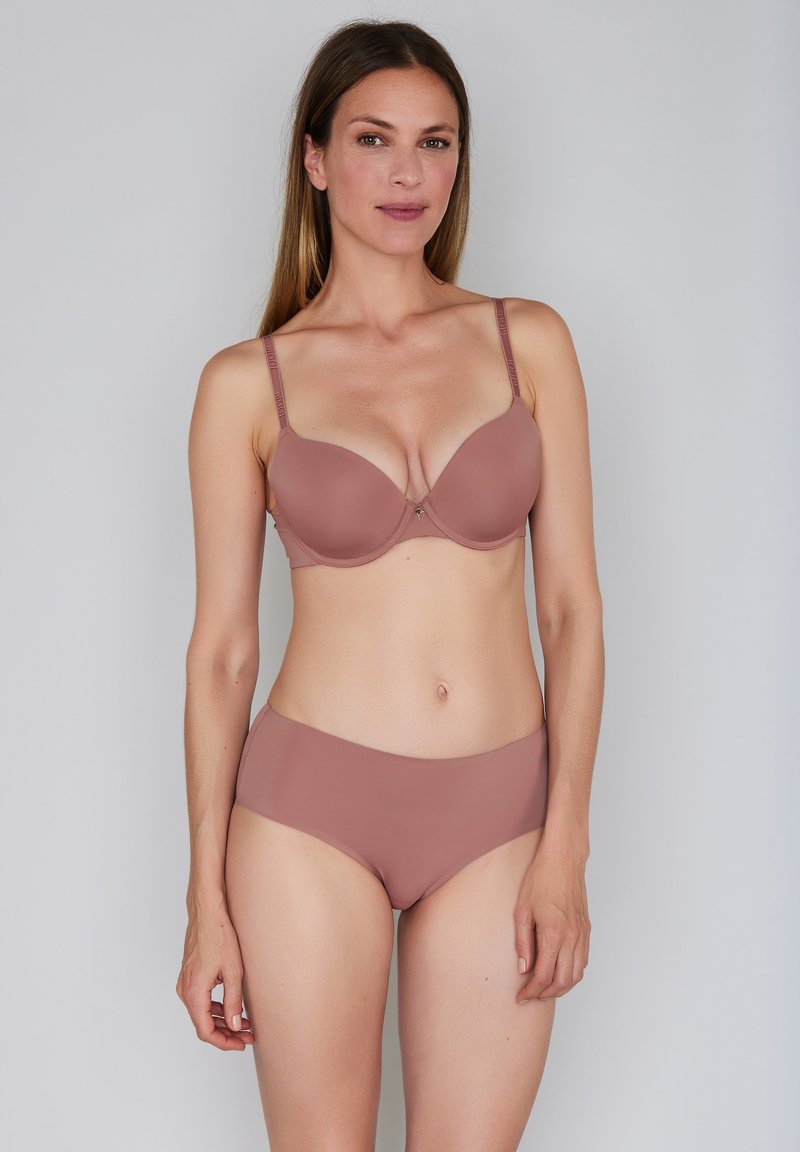 Nainen seisoo yksiväristä taustaa vasten, yllään yhteensopiva vaaleanlila toppauksella varustettu rintaliivit ja hipsterityyliset alushousut, katsoo eteenpäin neutraali ilme kasvoillaan.