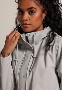 Light gray waterproof jacket with a high collar, adjustable drawstrings, button closure, and smooth texture, featuring a fitted silhouette.