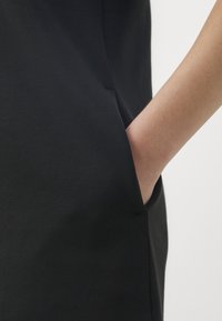 Main droite insérée dans la poche latérale d'un vêtement noir sur fond clair uni.