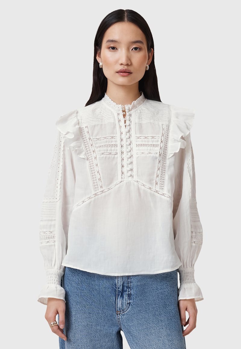 Blusa bianca con spalle arricciate, dettagli in pizzo intricati, una chiusura a bottoni e maniche lunghe, abbinata a jeans azzurri chiari.