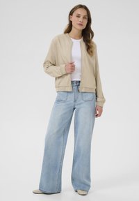 Veste bomber beige avec fermeture éclair à l'avant, poignets côtelés, associée à un pantalon en jean bleu clair à jambes larges. Le look est complété par des chaussures slip-on beiges.