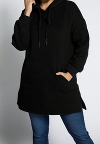 Sudadera negra larga hecha de material suave, con un bolsillo frontal, cordones ajustables y aberturas laterales para facilitar el movimiento.