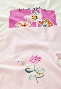 Zwei Baumwoll-T-Shirts gestapelt. Das obere hat ein pink-weißes Blumenmuster mit "BLOOM &". Das untere ist einfarbig hellrosa mit einem Blumenmuster.