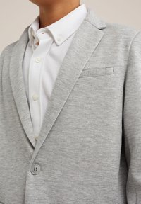 Kind, das ein weißes Hemd mit Knöpfen unter einem hellgrauen Ein-Knopf-Blazer trägt, der auf Höhe des Halses und der Mitte des Oberkörpers zugeschnitten ist.