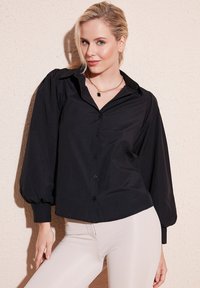 Black collared shirt with puffed long sleeves, button-up front, and a slightly cropped design, paired with fitted light-colored pants.