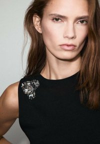 Top negro sin mangas que presenta un broche floral metálico con pétalos voluminosos y rizados y un acento central, destacando la textura y el diseño.