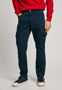 Pantalones cargo navy con un corte ajustado, que presentan bolsillos laterales y un cierre con botón. Combinados con zapatillas negras y un sudadera roja.