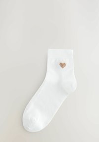 Vita bomullsankelsocka med ett mjukt broderat hjärta i tanfärg på utsidan. Har en ribbad mudd och en slät textur.