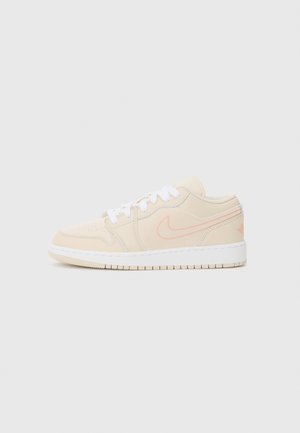 Beżowy niski sneaker z białymi sznurowadłami, różowym logo Nike i białą podeszwą na jednolitym jasnym tle.