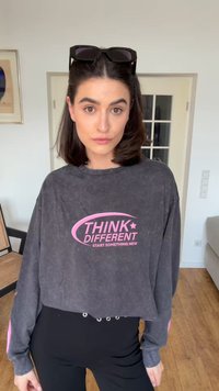 Νεαρή γυναίκα με σκουρόχρωμα μαλλιά φοράει μαύρα γυαλιά ηλίου στο κεφάλι και γκρι κοντό φούτερ με ροζ κείμενο «Think Different» σε εσωτερικό χώρο.