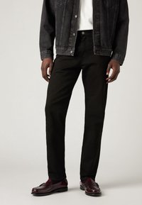 Homme portant un jean noir, une veste en jean noire, une chemise blanche et des mocassins bordeaux, debout devant un fond uni.
