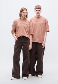 Δύο μοντέλα φορούν άνετες, κοντομάνικες μπλούζες σε χρώμα σκονισμένο ροζ με την επιγραφή «BROKE SINCE MILANO», συνδυασμένες με σκούρα καφέ παντελόνια με φαρδιά μπατζάκια.