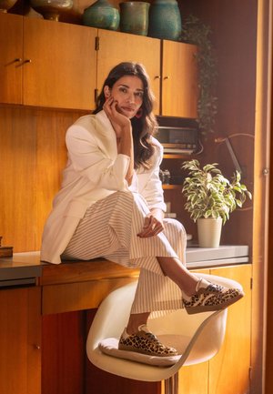 Luipaardprint sneakers met beige zolen, gedragen met een witte blazer en gestreepte broeken, zittend op een stoel bij een houten balie.