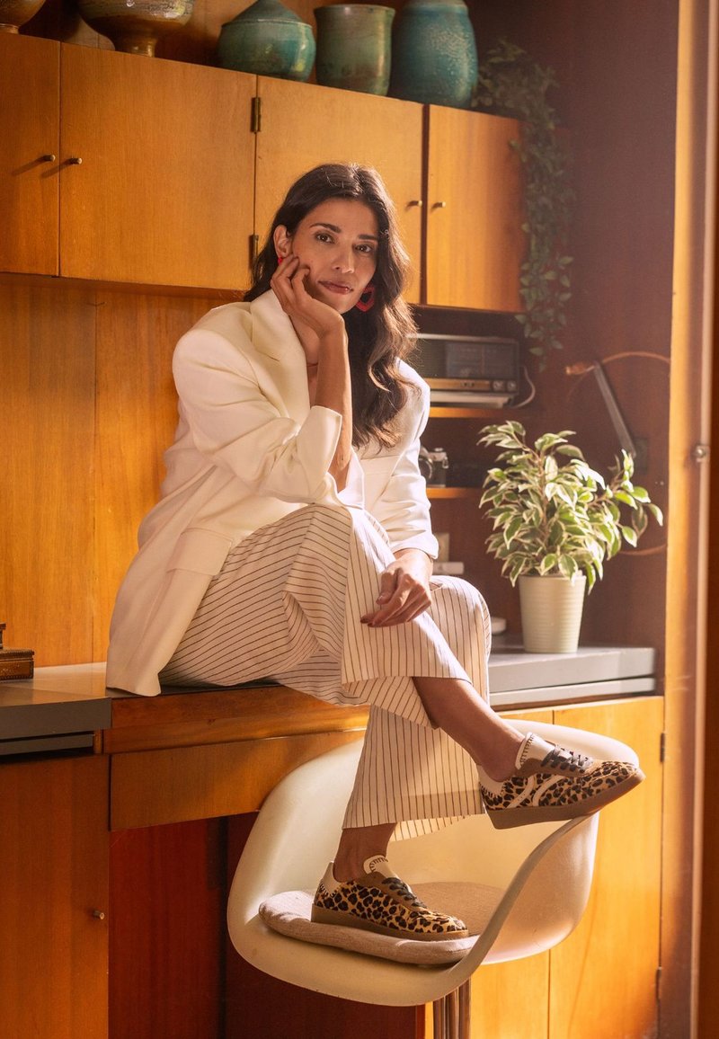 Luipaardprint sneakers met beige zolen, gedragen met een witte blazer en gestreepte broeken, zittend op een stoel bij een houten balie.