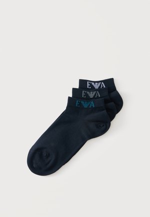 Trois paires de chaussettes noires montantes empilées, chacune avec un logo et un texte de couleur différente près du bord-côte.