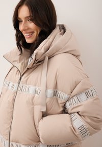 Uśmiechnięta kobieta w beżowej kurtce puchowej z kapturem, z detalami w postaci białej wstążki z logo, stojąca na tle gładkiej ściany.