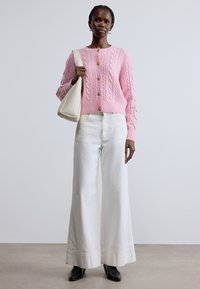 Rózsaszín kötött kardigán arany gombokkal, fehér bő szárú nadrággal és fekete magassarkú csizmával párosítva, krémszínű válltáskát tartva.