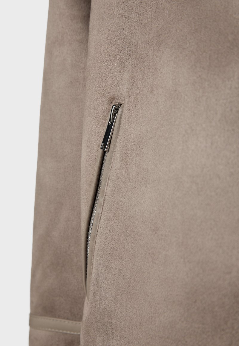 Manga de chaqueta de ante beige con un detalle de bolsillo con cremallera plateada vertical y un sutil dobladillo cosido cerca del puño.