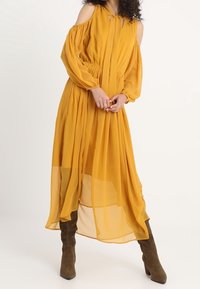 Musztardowa sukienka bez ramiączek z długimi rękawami, z marszczoną talią, zwiewnym materiałem z chiffonu oraz przezroczystą wstawką u dołu, zestawiona z brązowymi botkami.