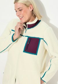 Kurtka polarowa w kremowym kolorze z akcentami w kolorze teal i burgundowym, z zamykaną na zamek kieszenią na klatce piersiowej oraz kieszeniami bocznymi, wysokim kołnierzem i zamkiem z przodu.