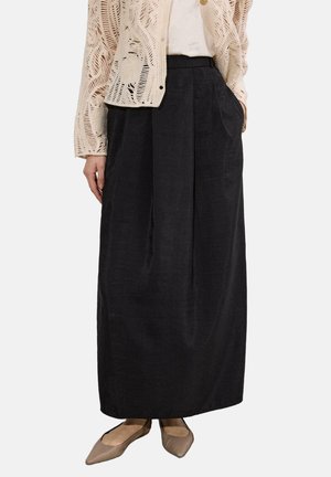 Γυναίκα που φοράει μπεζ δαντελένια ζακέτα, λευκό τοπ, μαύρη μακριά φούστα με τσέπες και μπεζ μυτερές μπαλαρίνες, στέκεται σε λευκό φόντο.