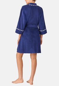 Donna in piedi a piedi nudi che indossa un accappatoio blu navy a maniche corte con rifiniture bianche ondulate su polsini e colletto, vista da dietro.