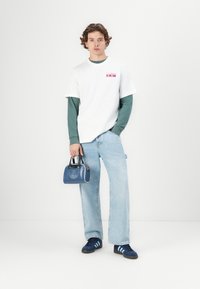 Hvit t-skjorte lagdelt over teal langermet skjorte, lyseblå vide jeans, marineblå sneakers med tre striper, og en liten blå håndveske med logo.