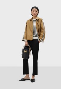 Braune verkürzte Jacke mit weißen und schwarzen Streifen, gepunkteten Bündchen; gestreiftes Oberteil, schwarze Hose, schwarze Handtasche mit goldenen Akzenten, flache schwarze Schuhe.