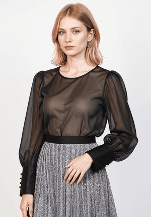 Joven mujer con cabello rubio hasta los hombros, vistiendo una blusa negra transparente y una falda plateada texturizada, de pie contra un fondo claro y liso.