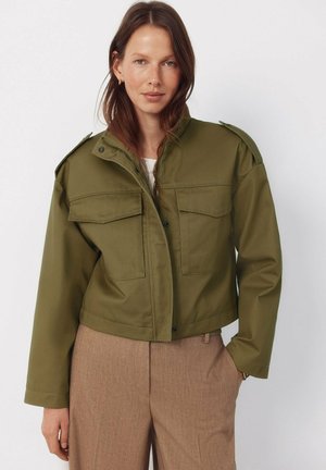 Kobieta ubrana w krótki oliwkowozielony żakiet z klapkowymi kieszeniami, połączony z wysokim stanem beżowych spodni, stojąca na tle białego, gładkiego tła.