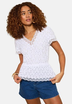 Moteris su garbanotais plaukais, dėvinti baltą trumpomis rankovėmis palaidinę su nėrinių apvadėliais ir mėlynas džinsines šortus, žvelgianti į šoną.