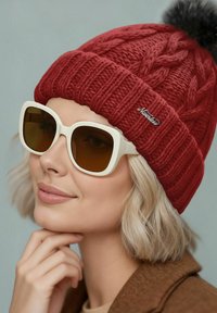 Red cable-knit beanie with a pom-pom on top and a small logo. Worn with oversized, white frame sunglasses and a brown coat.