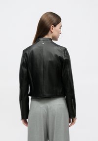 Casaco de couro preto com design ajustado e gola alta, apresentando punhos com fecho de correr e textura suave, visto pelas costas.